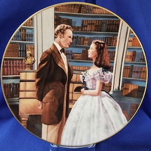 May include: A decorative plate featuring a scene with a man and woman in a library setting. The man wears a brown suit, and the woman is in a white dress. The plate has a gold rim and is displayed on a clear stand.