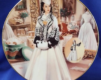 Peut inclure: Assiette décorative avec un bord doré, représentant une femme en robe blanche avec des détails noirs et blancs, un chapeau blanc et un petit sac à main blanc. L'assiette montre une scène de film.