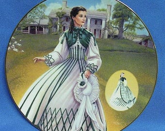 Peut inclure: Assiette décorative avec une femme en robe blanche et verte devant une grande maison. Le bord de l'assiette est doré. Une petite image de la femme est également présente. Fond vert et ciel bleu.