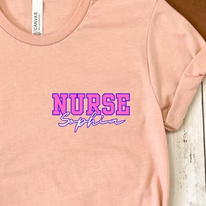 Peut inclure: Un t-shirt pêche avec des manches retroussées. Le t-shirt présente le texte "NURSE Sophia" dans un design violet et bleu sur le haut de la poitrine gauche. Le t-shirt est fait d'une matière douce et a une coupe décontractée.
