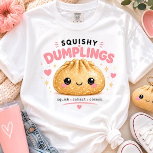 May include: White t-shirt with the text "SQUISHY DUMPLINGS" and a cartoon dumpling illustration. The design includes pink hearts and stars. A pink tumbler with a heart design, a pink scrunchie, and white sneakers are also visible.