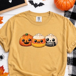 May include: A mustard yellow t-shirt featuring a graphic print of three cute, round characters: a smiling jack-o'-lantern pumpkin, a candy corn piece with a happy face, and a small skull with a skeleton body. The shirt has a Comfort Colors label.