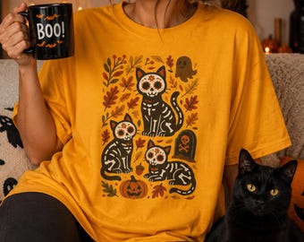 May include: A mustard yellow t-shirt featuring a graphic print of three black cats with white skeleton patterns, surrounded by autumn leaves, a pumpkin, a ghost, and a tombstone. A black mug with the text BOO and small bat icons is held.