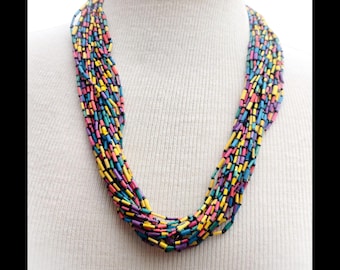 May include: Multicolored beaded necklace with black accents. The necklace is made of small, cylindrical beads in shades of yellow, pink, blue, and green. The beads are strung together on a black thread.
