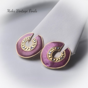 May include: Pair of vintage earrings with a C-shaped design. The earrings feature a gold-tone border with a purple enamel center. The center has a decorative gold and blue pattern. The text "Mohr Vintage Finds" is in the upper left corner.
