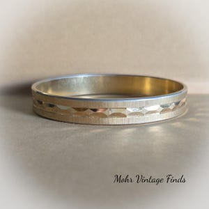 May include: A gold-toned bangle bracelet with a textured design. The bracelet is made of metal and has a smooth, polished finish.
