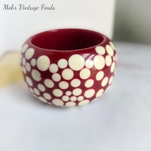 May include: A thick, rounded bangle bracelet featuring a deep red base color decorated with a pattern of cream-colored circular dots of varying sizes scattered across the entire exterior surface. The text Mohr Vintage Finds is visible.