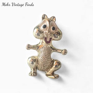 May include: A gold-toned squirrel brooch with black eyes and a red mouth. The squirrel is in a playful pose with its arms outstretched. The brooch has a textured body and is set against a white background. The text "Mohr Vintage Finds" is in the upper left corner.