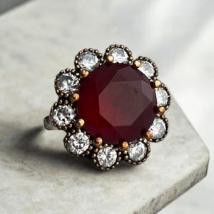 May include: A silver ring with a large, dark red gemstone surrounded by smaller, clear gemstones. The ring has a floral design.
