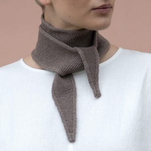 Pode incluir: Um lenço de pescoço tricotado na cor taupe com textura de ponto musgo. O cachecol está atado com um nó simples na frente, com duas pontas afiadas penduradas. É usado sobre uma camisa branca de manga comprida com decote redondo.