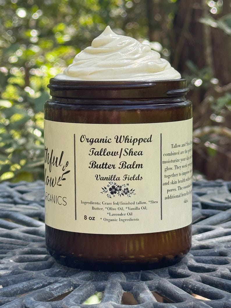 May include: A brown glass jar with a white label that reads "Organic Whipped Tallow/Shea Butter Balm Vanilla Fields". The jar contains a white, whipped cream-like substance. The label also lists the ingredients and size: 8 oz. Organic Ingredients.