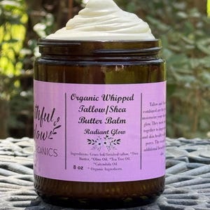 May include: A glass jar with a purple label that reads "Organic Whipped Tallow/Shea Butter Balm Radiant Glow" and lists the ingredients: "Grass fed/finished tallow, Shea Butter, Olive Oil, Tea Tree Oil, Calendula Oil". The jar contains a white whipped balm and is labeled "8 oz * Organic Ingredients".