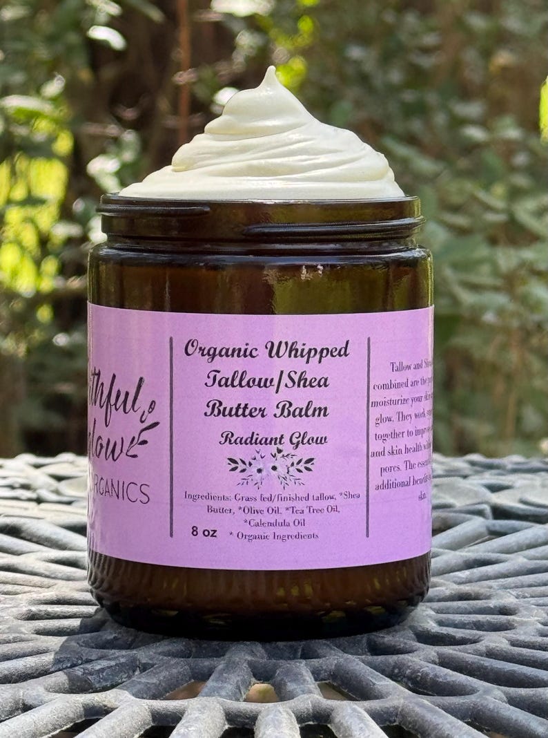 May include: A glass jar with a purple label that reads "Organic Whipped Tallow/Shea Butter Balm Radiant Glow" and lists the ingredients: "Grass fed/finished tallow, Shea Butter, Olive Oil, Tea Tree Oil, Calendula Oil". The jar contains a white whipped balm and is labeled "8 oz * Organic Ingredients".