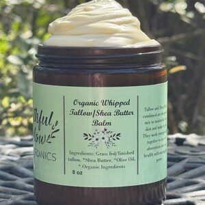 May include: A glass jar with a green label that reads "Organic Whipped Tallow/Shea Butter Balm". The label also lists the ingredients: Grass fed/finished tallow, *Shea Butter, *Olive Oil, *Organic Ingredients. The jar contains a white whipped balm and is sitting on a gray surface.