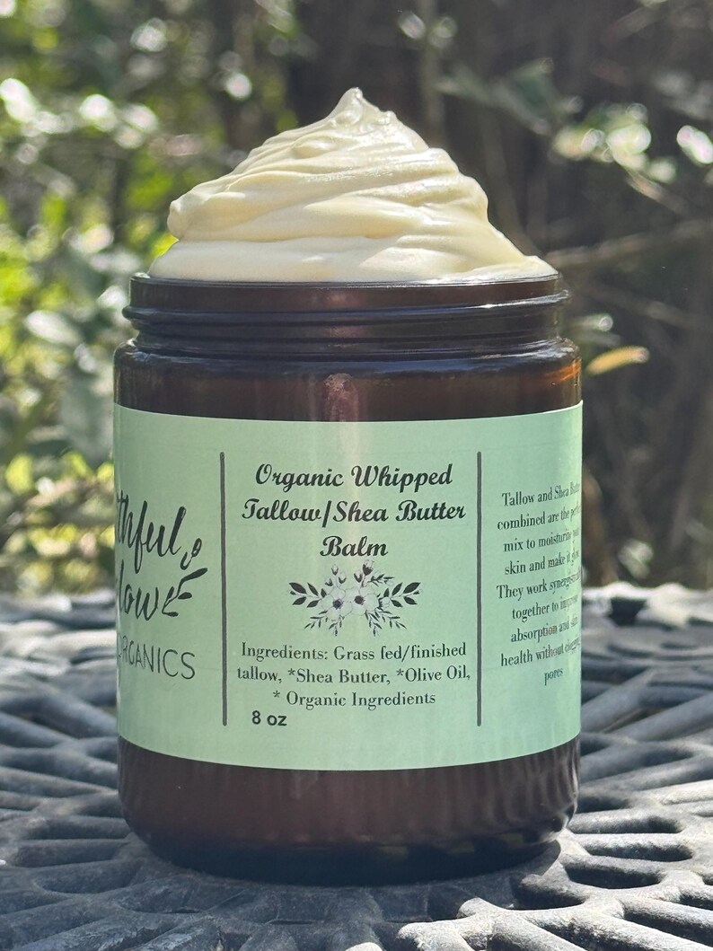 May include: A glass jar with a green label that reads "Organic Whipped Tallow/Shea Butter Balm". The label also lists the ingredients: Grass fed/finished tallow, *Shea Butter, *Olive Oil, *Organic Ingredients. The jar contains a white whipped balm and is sitting on a gray surface.