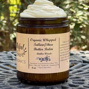 May include: A brown glass jar with a white label that reads "Organic Whipped Tallow/Shea Butter Balm Amber Woods" and lists the ingredients: "Grass fed/finished tallow, Shea Butter, Olive Oil, Vanilla Oil, Sandalwood Oil". The jar contains a white, whipped balm and is labeled "8 oz *Organic Ingredients".