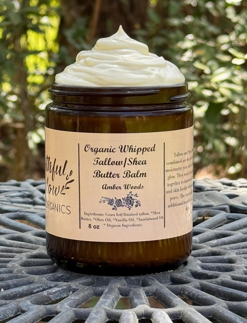 May include: A brown glass jar with a white label that reads "Organic Whipped Tallow/Shea Butter Balm Amber Woods" and lists the ingredients: "Grass fed/finished tallow, Shea Butter, Olive Oil, Vanilla Oil, Sandalwood Oil". The jar contains a white, whipped balm and is labeled "8 oz *Organic Ingredients".