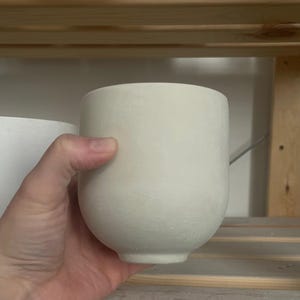 Może przedstawiać: Biała ceramiczna doniczka z zaokrąglonym dnem i gładką, fakturowaną powierzchnią.