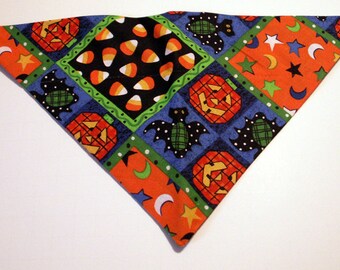 May include: A triangular pet bandana featuring a patchwork pattern of Halloween motifs including orange jack-o'-lanterns, black bats, and candy corn on black, orange, and blue backgrounds with green borders decorated with stars and crescent moons.