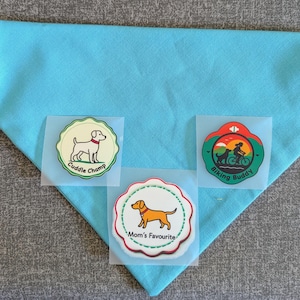 Puede incluir: Un pañuelo azul claro para perros con tres parches termoadhesivos. Los parches presentan perros de dibujos animados y las frases "Cuddle Champ", "Biking Buddy" y "Mom's Favourite". El pañuelo tiene forma triangular.