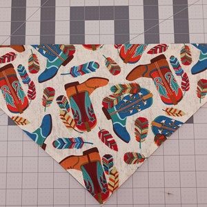 May include: A triangular pet bandana featuring a repeating pattern of cowboy boots in shades of orange, blue, and red, interspersed with colorful feathers on a light gray background with a subtle heathered texture throughout the fabric.