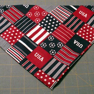 Può includere: Una bandana triangolare con un design patchwork di quadrati di tessuto rossi, bianchi e blu. Il motivo include blocchi alternati di stelle bianche su sfondo blu, strisce rosse e bianche e quadrati rossi con il testo bianco USA stampato.