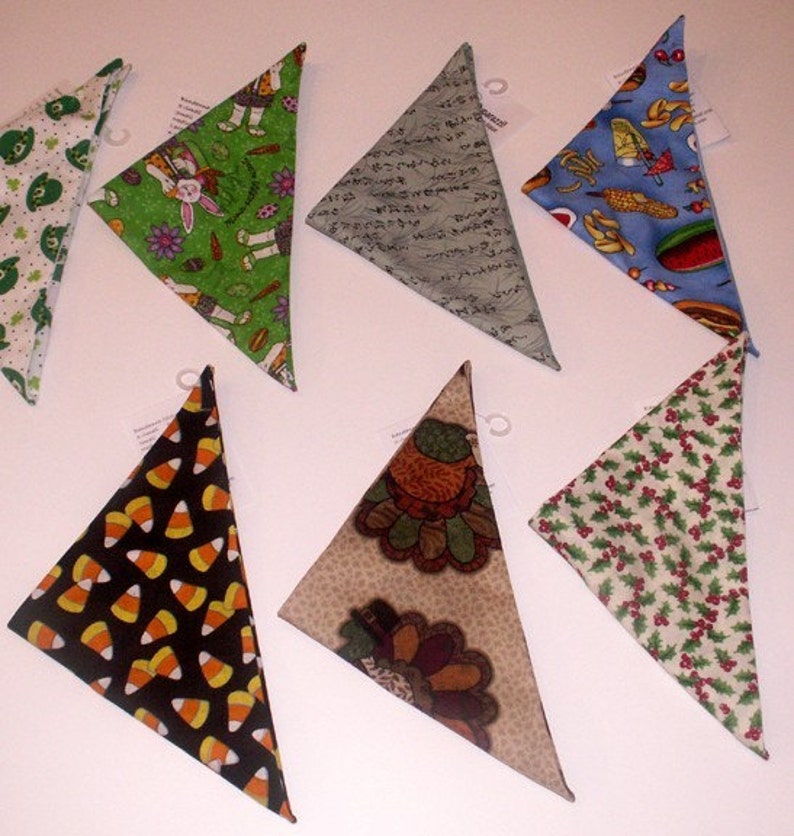 May include: A collection of triangular bandanas with various patterns. Designs include candy corn on black, a turkey, green shamrocks, a bunny with carrots, Japanese script, and a food-themed pattern with hot dogs. The bandanas are likely for pets.