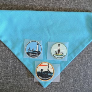 Puede incluir: Un pañuelo turquesa para mascotas con tres parches circulares. Los parches presentan ilustraciones de faros con el texto "Ocracoke Lighhouse", "Ocracoke Island" y "Cape Hatteras". Los parches tienen un borde blanco.