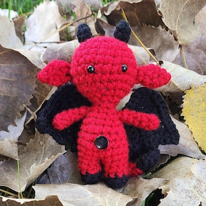 May include: A small red crocheted devil with black horns and wings. The devil is sitting on a bed of brown leaves.