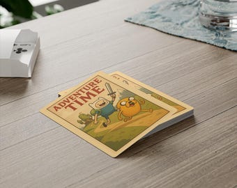 Op de afbeelding: Een kaartspel van Adventure Time met Finn en Jake. De kaarten hebben een beige achtergrond met het logo van de show en illustraties van personages. Het spel ligt op een houten tafel.