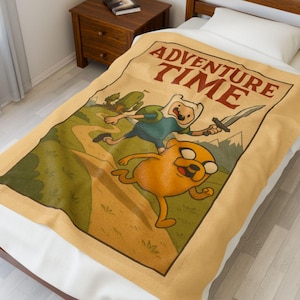 May include: A tan Adventure Time blanket featuring Finn and Jake running through a landscape. The blanket has the words "Adventure Time" in red letters at the top. The background has green hills and mountains.