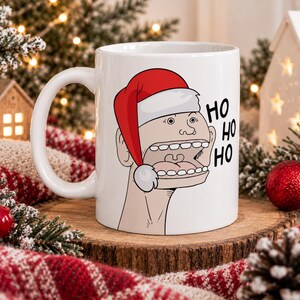 May include: A white ceramic mug featuring a graphic illustration of a stylized face wearing a red and white Santa hat with its mouth wide open, accompanied by the black text HO HO HO printed vertically on the right side of the design.