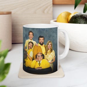 May include: White ceramic mug with a colorful image of a group of people wearing yellow clothing. The image includes a man with a beard and glasses, a woman with long blonde hair, and a man with a black cat. The text "The Gang" is visible on the mug.