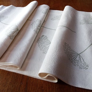 May include: A set of four white linen table runners with a green floral design. The runners are folded and stacked on a wooden surface.