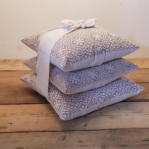 May include: Three decorative throw pillows with a light purple and white geometric pattern. The pillows are stacked on top of each other and tied together with a white ribbon.