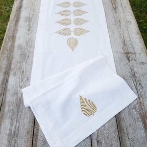 May include: White table runner with a gold leaf design. The runner is folded in half and has a single leaf design on the folded side.