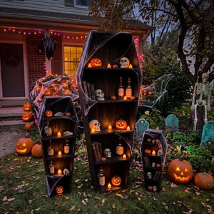 May include: Three black coffin-shaped shelves of varying heights arranged outdoors, decorated with artificial spider webs, human skulls, glass bottles, and small glowing orange LED tea lights on each shelf level.