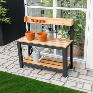May include: A black metal garden work bench with a wooden top and shelves. The bench has two terracotta pots on the top shelf and a watering can on the lower shelf. The bench is in front of a window with plants visible outside.