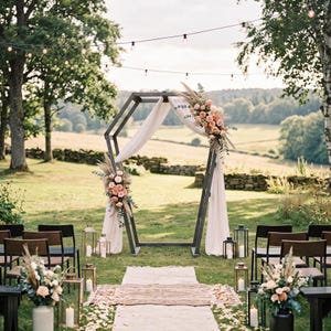 May include: A dark metal hexagonal wedding arch draped with white fabric and decorated with peach and pink floral arrangements. The setup includes a white aisle runner, metal lanterns with candles, and wooden chairs on a grassy field.