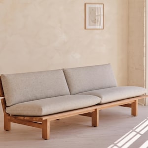 May include: A long sofa featuring a natural light brown wooden frame with a slatted back design and two thick, light gray upholstered seat cushions and two matching back cushions, supported by six sturdy wooden legs in a low-profile structure.