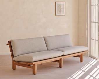 May include: A long sofa featuring a natural light brown wooden frame with a slatted back design and two thick, light gray upholstered seat cushions and two matching back cushions, supported by six sturdy wooden legs in a low-profile structure.