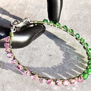 May include: A beaded bracelet featuring a gradient of pink and green faceted beads. The beads are strung on a wire with a silver clasp. The wire is wrapped with green wire. The bracelet is displayed on a black surface.