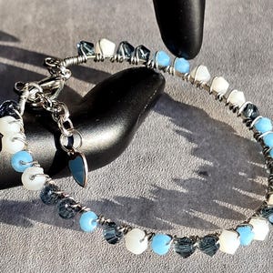 May include: A beaded bracelet featuring a mix of white, light blue, and dark blue beads strung on a silver wire. The bracelet has a toggle clasp and a small, blue heart-shaped charm. The beads are arranged in a pattern around the wrist.