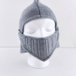 May include: Gray fleece knight helmet with a pointed top and a face mask. The helmet is on a white mannequin head.