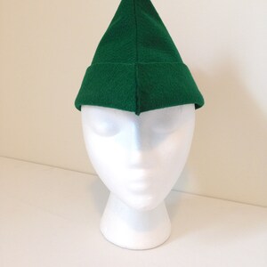 May include: A green felt elf hat with a pointed tip. The hat is on a white mannequin head.