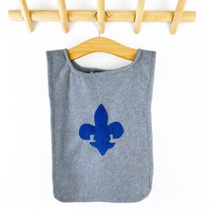 May include: Gray fleece costume vest with a blue fleur-de-lis emblem. The vest is hanging on a wooden coat rack.