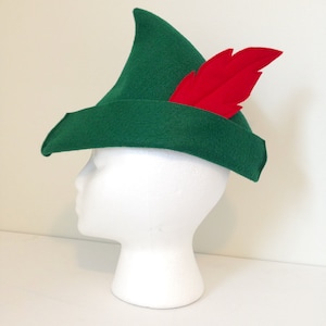 May include: A green felt hat with a pointed crown and a red feather. The hat is on a white mannequin head.
