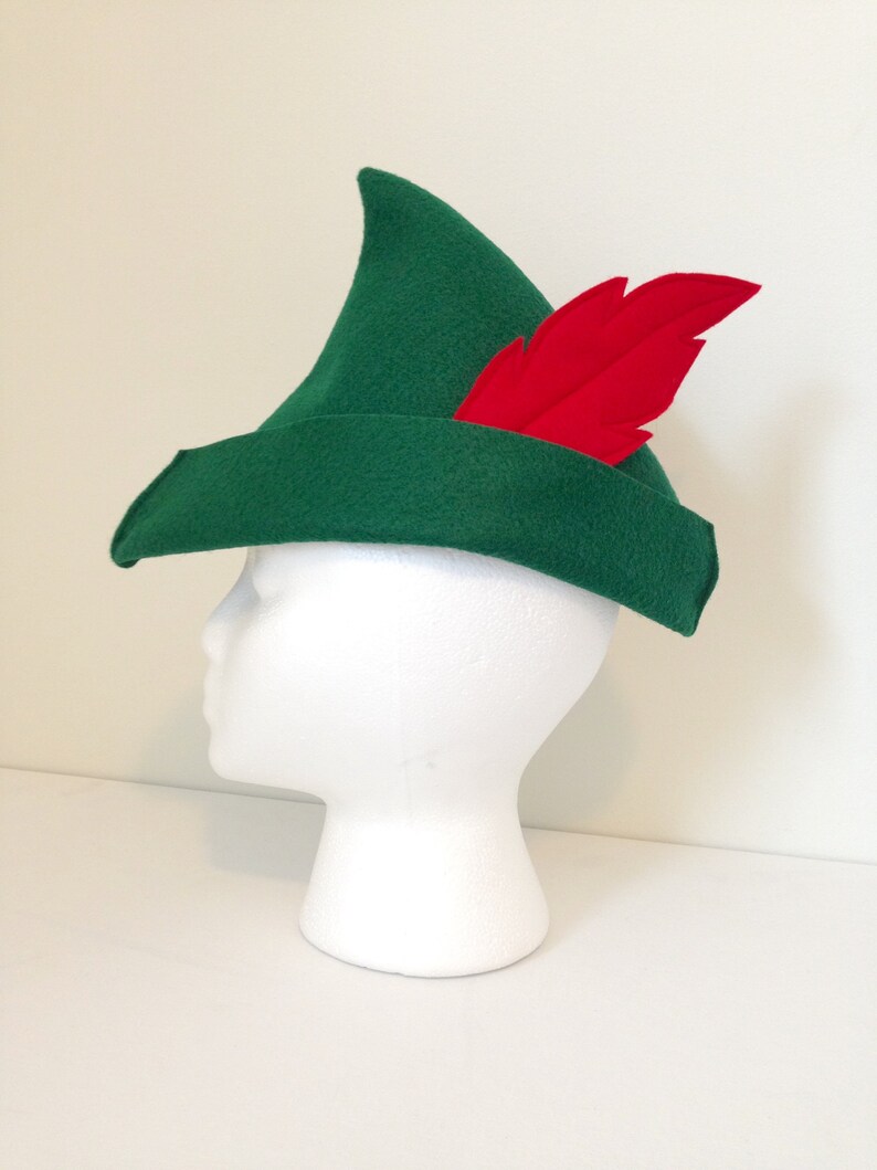 May include: A green felt hat with a pointed crown and a red feather. The hat is on a white mannequin head.