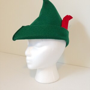 May include: A green felt hat with a pointed tip and a red feather. The hat is on a white mannequin head.