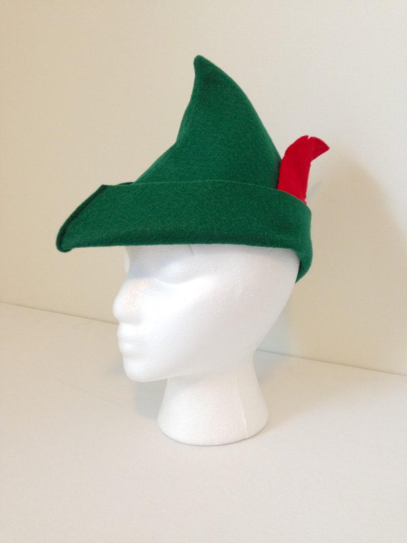 May include: A green felt hat with a pointed tip and a red feather. The hat is on a white mannequin head.